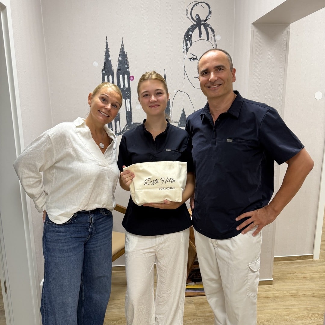 Auszubildende Klara steht zwischen zwei Teammitgliedern der Zahnarztpraxis und hält eine Stofftasche mit der Aufschrift „Erste Hilfe für Azubis".